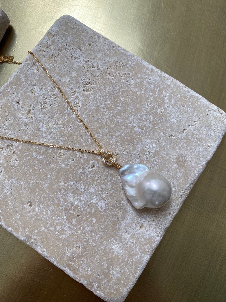 Pu&ograve; includere: Una collana con catena dorata con un grande ciondolo a forma di perla barocca bianca. Il ciondolo &egrave; attaccato alla catena con un piccolo anello dorato che &egrave; incastonato con piccole pietre trasparenti.