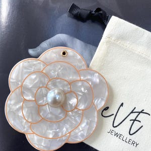 Op de afbeelding: Een grote, bloemvormige hanger met een parelmoer afwerking en een centrale parel. De hanger heeft een goudkleurige rand en een klein gaatje aan de bovenkant. Een klein zakje met trekkoord met de tekst "eve JEWELLERY" staat ook op de afbeelding.