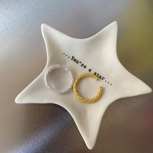 Op de afbeelding: Twee textuurrijke metalen ringen, een zilveren en een gouden, worden gepresenteerd op een wit stervormig schaaltje. Het schaaltje heeft de tekst "...You're a star..." erop gedrukt.