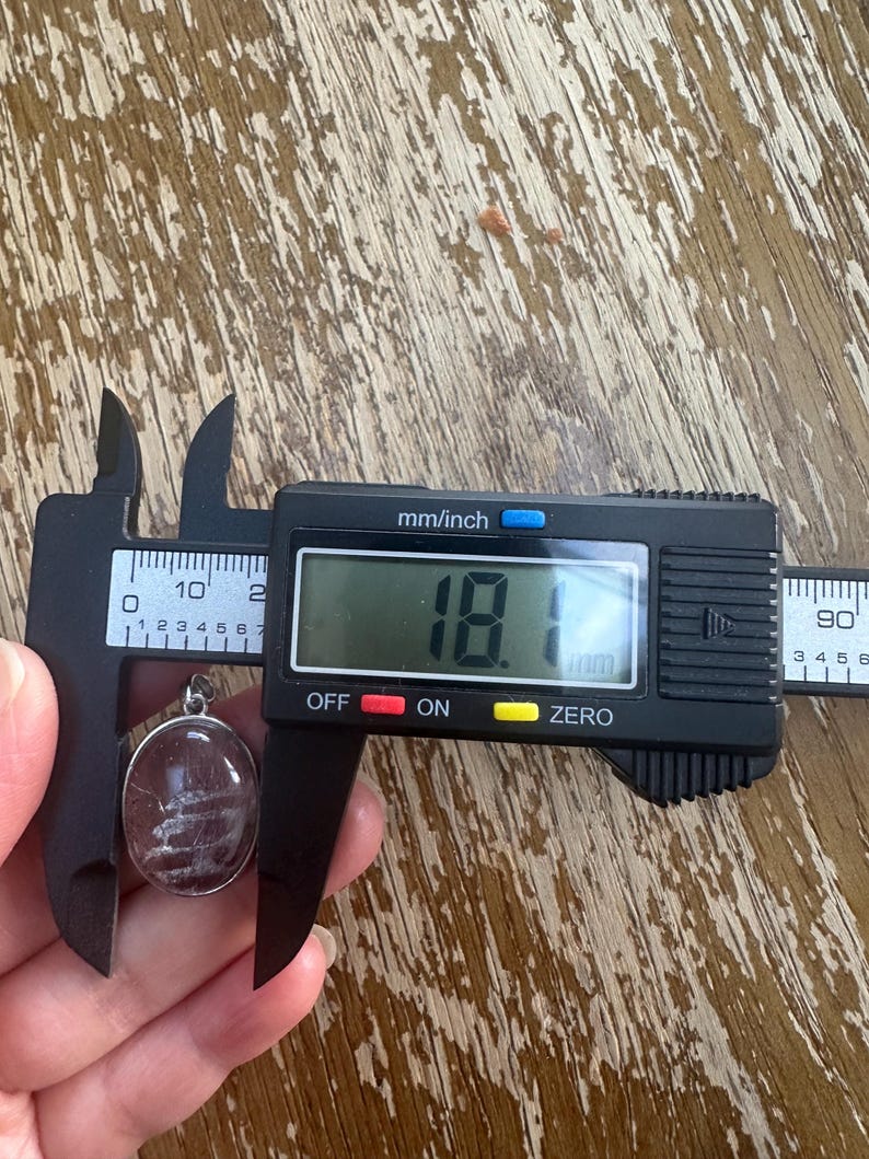 Op de afbeelding: Een ovale hanger met een helder middenstuk en zilverkleurige metalen rand wordt gemeten met een digitale schuifmaat. De hanger heeft witte, lineaire insluitsels. De schuifmaat geeft 1,81 cm aan.