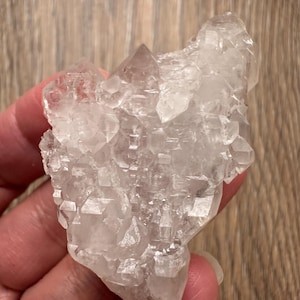 Puede incluir: Un grupo de cristales de cuarzo transparente con múltiples puntas y una superficie rugosa y texturizada. El cristal se sostiene en una mano, mostrando su forma natural e irregular y su calidad translúcida. El cuarzo tiene un aspecto claro, casi incoloro.