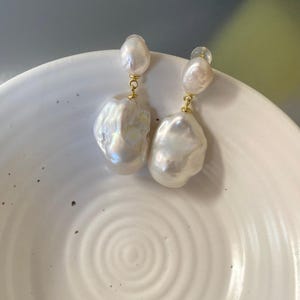 May include: A pair of elegant pearl earrings. Each earring features a small, round pearl stud connected to a larger, baroque-shaped pearl. The pearls have a lustrous, iridescent sheen and are suspended by gold-colored metal findings.