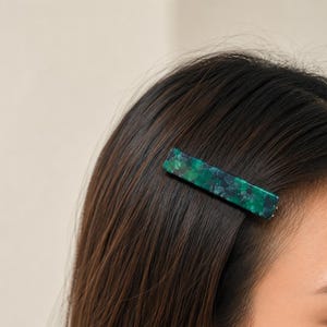 Puede incluir: Una pinza rectangular para el cabello con un patr&oacute;n verde y azul veteado est&aacute; sujeta al cabello casta&ntilde;o oscuro. La pinza est&aacute; hecha de un material brillante y est&aacute; colocada cerca de la parte superior de la cabeza. El fondo es de color neutro.