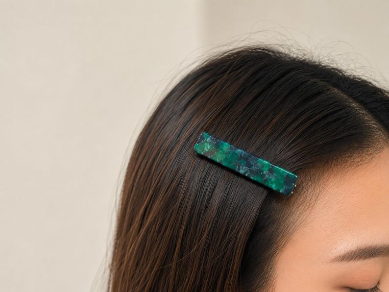 Puede incluir: Una pinza rectangular para el cabello con un patr&oacute;n verde y azul veteado est&aacute; sujeta al cabello casta&ntilde;o oscuro. La pinza est&aacute; hecha de un material brillante y est&aacute; colocada cerca de la parte superior de la cabeza. El fondo es de color neutro.