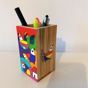 May include: A wooden pencil holder with a colorful painted design featuring birds, a rabbit, and a house. The holder is filled with pens and markers.