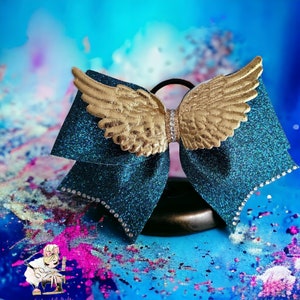 Könnte beinhalten: Eine tealblaue Glitzer Schleife mit goldenen Flügeln und Strasssteinen. Die Schleife ist mit einem schwarzen Gummiband gebunden.