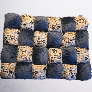 Könnte beinhalten: Eine Patchwork-Hundecke mit abwechselnden marineblauen und gemusterten Quadraten. Die gemusterten Quadrate zeigen Pfotenabdrücke, Knochen und das Wort "Woof" in Braun, Beige und Rot. Die Decke ist rechteckig und wirkt weich.