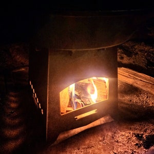 Puede incluir: Una pequeña estufa de leña de metal con fuego ardiendo en su interior. La estufa está sentada sobre una superficie de tierra.