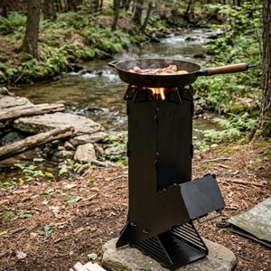 Può includere: Un fornello da campeggio in metallo nero con una padella che cucina cibo su una fiamma libera. Il fornello è posizionato su una roccia in un ambiente forestale, con un ruscello sullo sfondo. Legna da ardere è impilata nelle vicinanze.