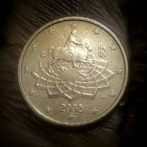 Puede incluir: Una moneda de 1 euro de color dorado con la imagen de una mujer montando a caballo sobre un mapa. La moneda tiene 12 estrellas alrededor del borde y el año 2002 impreso en la parte inferior.