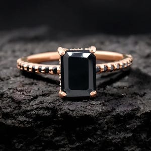 May include: A rose gold ring featuring a large, rectangular black gemstone. The band is embellished with small black stones. The ring is set against a dark, textured surface.