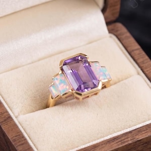 May include: A gold ring featuring a large rectangular purple gemstone, flanked by two smaller opal stones. The ring is presented in a wooden jewellery box with a cream-coloured interior. The gemstones are set in a gold band.
