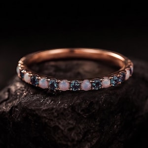 May include: Rose gold ring with alternating opal and blue gemstones. The ring is set on a dark, textured surface, showcasing the delicate details of the jewelry. The gemstones are evenly spaced around the band.