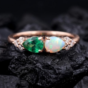 May include: Rose gold ring featuring a pear-shaped emerald and a pear-shaped opal. The ring is accented with small diamonds on either side of the gemstones. The ring is set against a dark background.