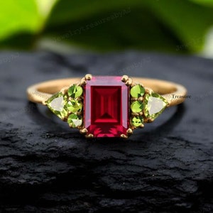 May include: A gold ring with a large, square, red gemstone surrounded by smaller, green gemstones. The ring is set on a black background.