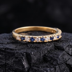 Könnte beinhalten: Ein goldener Bandring mit abwechselnd blauen und weißen Edelsteinen. Der Ring liegt auf einer dunklen, strukturierten Oberfläche, die den Kontrast zwischen dem glänzenden Gold und den Edelsteinen hervorhebt. Die Edelsteine sind gleichmäßig angeordnet.