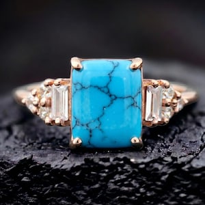 May include: A rose gold ring with a rectangular turquoise stone, displaying black veining. The ring is set with two baguette-cut diamonds and smaller accent stones. The ring is set against a dark, textured background.