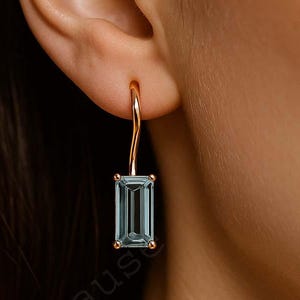 May include: A rose gold earring featuring a rectangular, pale blue gemstone. The gemstone is set in a simple design. The earring is suspended from the ear.