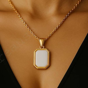 Könnte beinhalten: Eine goldfarbene Halskette mit einem rechteckigen Anhänger. Der Anhänger hat eine weiße Mitte und einen goldenen Rand. Die Kette ist ebenfalls goldfarben und hat ein strukturiertes Design. Die Halskette wird von einer Person mit einem schwarzen Oberteil getragen.