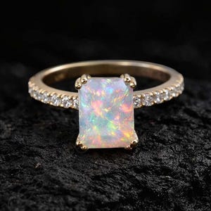 May include: A gold ring with a rectangular opal gemstone as the centerpiece. The band is adorned with small, clear gemstones. The opal displays a play of colors, including white, yellow, and green, set against a dark background.