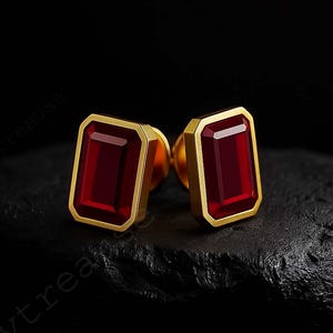 May include: A pair of gold-toned stud earrings with rectangular, deep red gemstones. The gemstones are set within a gold frame, creating a bold and elegant look. The earrings are displayed on a dark, textured surface.