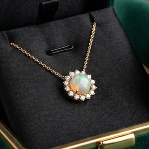 Könnte beinhalten: Eine goldene Halskette mit einem Opalanhänger, umgeben von kleinen, klaren Edelsteinen. Präsentiert in einer offenen, grünen Samt-Schmuckschatulle mit dem Aufdruck "ELEGANCE".