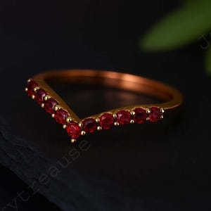 Puede incluir: Un anillo dorado en forma de V adornado con gemas rojas brillantes. El diseño del anillo presenta una fila de piedras rojas de corte redondo a lo largo de la forma de V, creando un contraste llamativo con la banda dorada. El anillo se muestra sobre una superficie oscura.