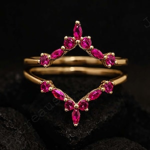 Könnte beinhalten: Ein goldfarbener Ring mit leuchtend rosa Edelsteinen. Der Ring hat ein Doppelband-Design, wobei das obere Band V-förmig ist und mit mehreren marquiseförmigen und runden Edelsteinen besetzt ist. Die Edelsteine sind in einem leuchtenden Rosaton gehalten.