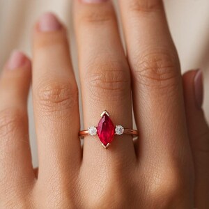 K&ouml;nnte beinhalten: Ein goldener Ring mit einem rotfarbenen, marquisef&ouml;rmigen Edelstein, flankiert von zwei kleineren, runden, klaren Edelsteinen. Der Ring ist auf einem zarten Goldband gefasst. Der Ring ist ein Schmuckst&uuml;ck.