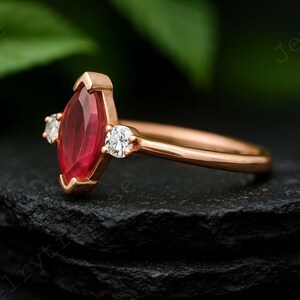 K&ouml;nnte beinhalten: Ein ros&eacute;goldener Ring mit einem markisef&ouml;rmigen Rubin-Mittelstein und zwei runden Diamant-Akzenten. Das Band und die Fassung des Rings haben eine warme, metallische Farbe, und die Edelsteine sind vor einem dunklen Hintergrund gesetzt.