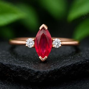 K&ouml;nnte beinhalten: Ein ros&eacute;goldener Ring mit einem tiefroten, marquisef&ouml;rmigen Edelstein im Zentrum, flankiert von zwei kleineren, runden, klaren Edelsteinen. Der Ring ist vor einem dunklen, strukturierten Hintergrund mit verschwommenem Gr&uuml;n platziert.