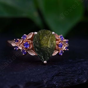 May include: A rose gold ring with a large, pear-shaped, olive-green gemstone at its centre. The ring is decorated with floral and leaf details, and small, round, blue gemstones. The ring is set against a dark background.