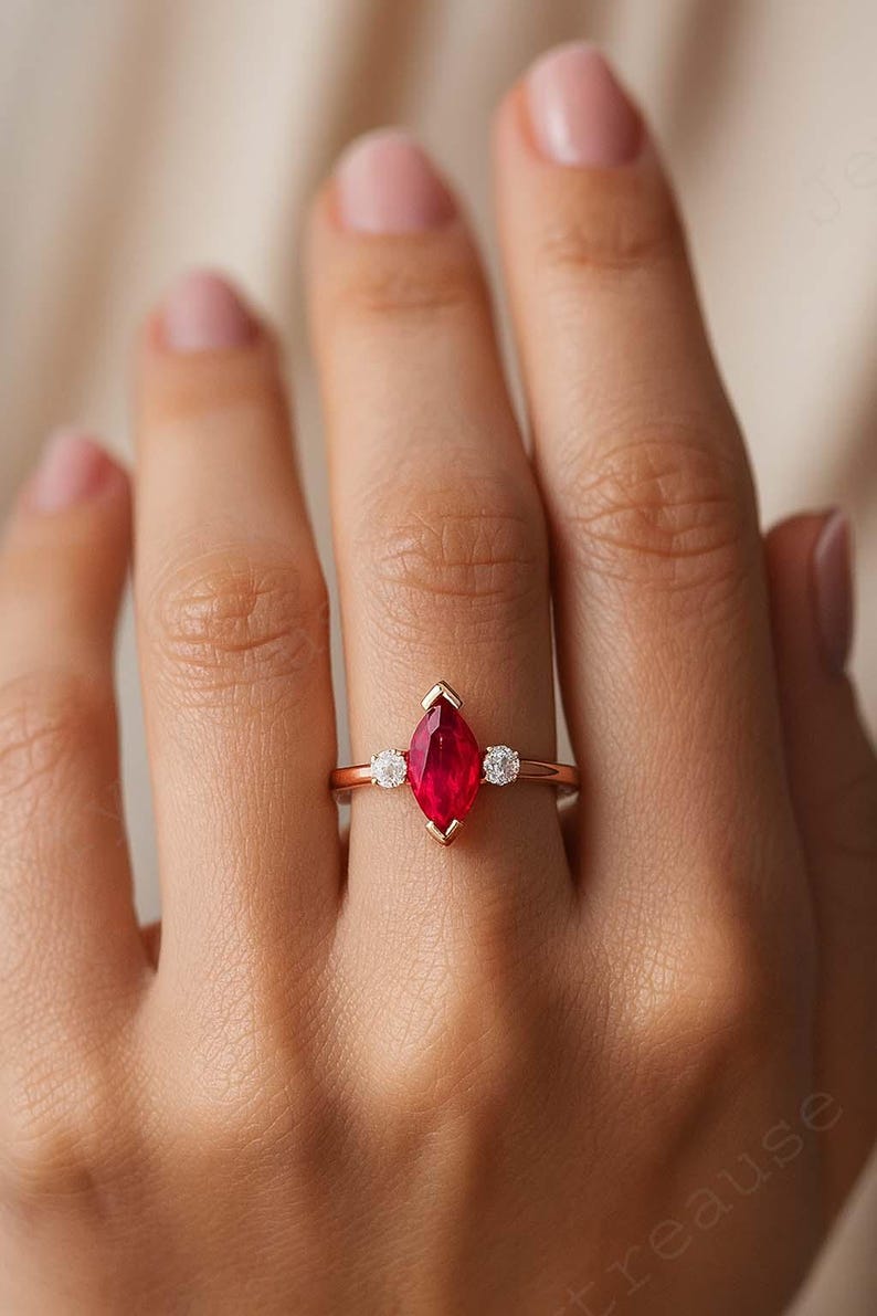 K&ouml;nnte beinhalten: Ein goldfarbener Ring mit einem markanten, marquisef&ouml;rmigen, leuchtend roten Edelstein im Mittelpunkt. Zwei kleinere, runde, klare Edelsteine flankieren den Hauptstein. Der Ring hat ein zartes Band und zeigt ein klassisches und elegantes Design.