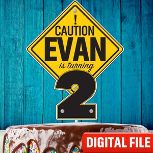 May include: Yellow and black caution sign with the text "CAUTION EVAN is turning 2" and a black number 2. The sign is on top of a chocolate cake with colorful sprinkles.