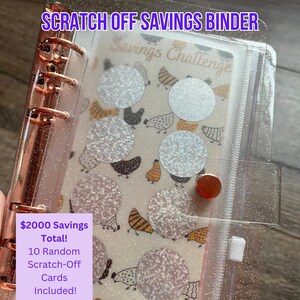 May include: A clear, glittery binder with rose gold hardware, labeled "SCRATCH OFF SAVINGS BINDER." The binder contains a savings challenge sheet with scratch-off circles and a chicken pattern. The text on the label reads "$2000 Savings Total! 10 Random Scratch-Off Cards Included!"