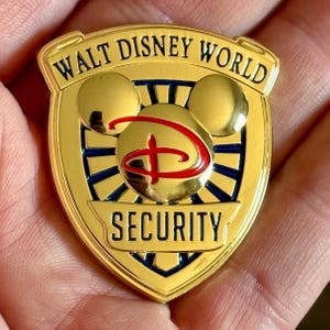 May include: A gold-toned metal shield-shaped pin featuring the text WALT DISNEY WORLD at the top and SECURITY at the bottom in black lettering. The center displays a gold Mickey Mouse head silhouette with a red stylized D logo over a blue sunburst.