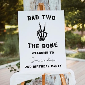 May include: A white sign with black text that reads "Bad Two The Bone Welcome to Jacob's 2nd Birthday Party". A black skeleton hand making a peace sign is above the text.
