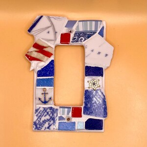 May include: A mosaic light switch cover with a nautical theme. The cover is made of broken ceramic pieces in shades of blue, white, and red. An anchor and a ship's wheel are included in the design. The background is a solid orange color.