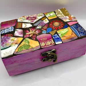 Könnte beinhalten: Eine rechteckige Holzschatulle in leuchtendem Violett mit einem Klappdeckel, der mit einem Mosaik aus bunten Keramikscherben verziert ist. Der Deckel zeigt verschiedene florale Muster, goldene Akzente und eine rote Blumenverzierung.