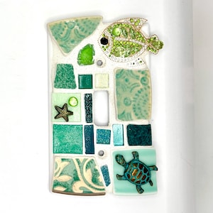 May include: A rectangular light switch cover plate decorated with a mosaic of green and teal ceramic tiles. The design features a jeweled green fish, a small starfish, and a turtle figurine embedded among the various textured tile pieces.