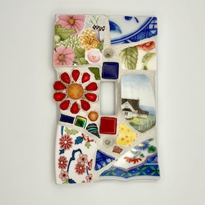 May include: A rectangular light switch cover plate featuring a mosaic design made of various ceramic shards. The pattern includes floral motifs, a small house illustration, a red flower-shaped piece, and blue and white geometric fragments.