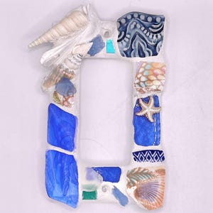 Puede incluir: Un marco de fotos rectangular decorado con conchas marinas, vidrio azul y mosaicos. El marco presenta una estrella de mar, varias conchas marinas y mosaicos azules y blancos. El marco es blanco con una abertura rectangular.