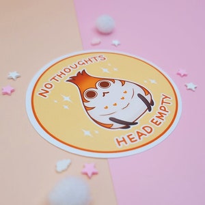 May include: A yellow round sticker with a cartoon character and the text "NO THOUGHTS HEAD EMPTY". The character is a white and orange creature with a round body and large eyes. The sticker is on a pink and beige background.