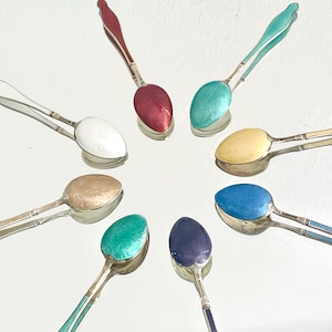 Könnte beinhalten: Eine Sammlung von acht kleinen Löffeln mit farbigen Emaille-Schalen und silbernen Griffen. Die Löffel haben verschiedene Farben, darunter Rot, Türkis, Gelb, Blau und Weiß. Die Löffel sind kreisförmig angeordnet.