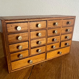 May include: A wooden apothecary cabinet with multiple small drawers. The cabinet is made of light brown wood and features brass knobs on each drawer. The drawers are arranged in rows and columns, creating a grid-like pattern.