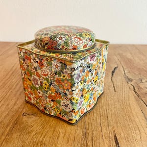 Könnte beinhalten: Eine vintage Blechdose mit floralem Muster und Deckel. Die Dose ist quadratisch mit abgerundeten Kanten und hat einen goldenen Rand. Das Blumenmuster ist bunt und zeigt eine Vielzahl von Blumen in den Farben Rosa, Gelb, Orange, Rot und Blau.
