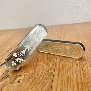 May include: Two rectangular, clear glass drawer pulls with etched floral designs. The pulls have a silver-coloured border and are resting on a wooden surface. The etched designs include a starburst and delicate sprigs with small, round accents.