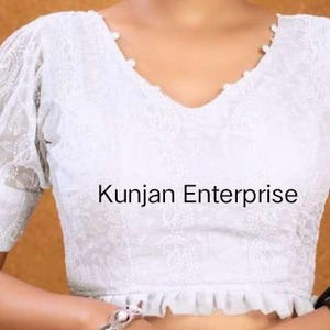 May include: White embroidered blouse with a V-neckline and puff sleeves. The blouse has a ruffled hem and is decorated with pearl-like buttons. The text "Kunjan Enterprise" is visible on the front of the blouse.