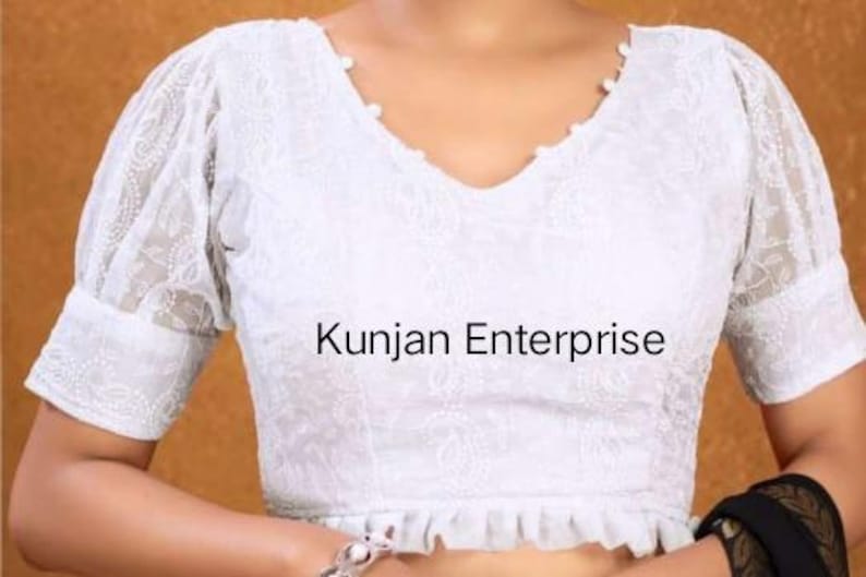May include: White embroidered blouse with a V-neckline and puff sleeves. The blouse has a ruffled hem and is decorated with pearl-like buttons. The text "Kunjan Enterprise" is visible on the front of the blouse.