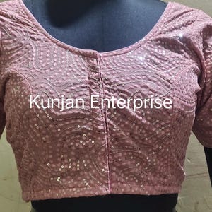 May include: A pink embroidered blouse with sequins and a button closure. The blouse has short sleeves and a round neckline. The text "Kunjan Enterprise" is visible on the front of the blouse.